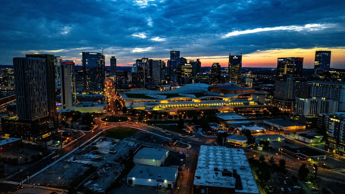 A vibrant city skyline illuminated at sunset, with buildings aglow and traffic lights capturing the dynamic energy of downtown Nashville. The scene conveys urban excitement and the convenience of nearby venues and nightlife.
