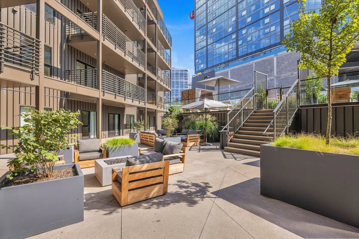 An inviting outdoor seating area featuring comfortable chairs, planters with greenery, and a staircase leading to an upper level, all shaded by a sunshade canopy. The scene hints at cozy gatherings, relaxation, and the potential for firepit lounges in cooler months.