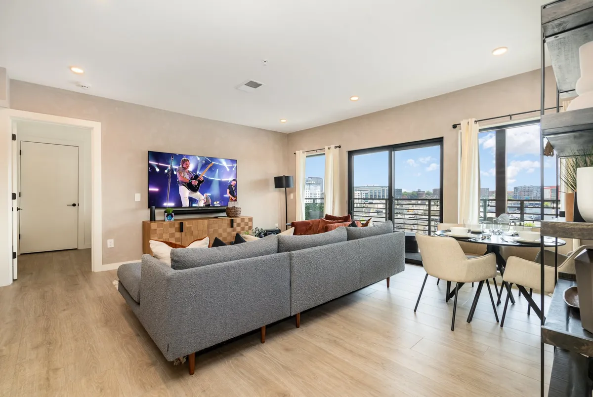 This image shows a large sectional sofa facing a television displaying a concert, with large windows that frame an urban cityscape beyond. The space feels cozy and social, with sunlight streaming in, ideal for movie nights or post-adventure relaxation.