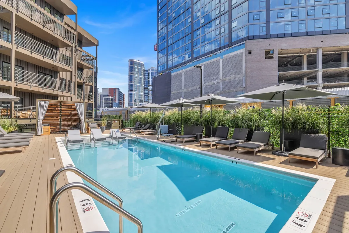 A vibrant swimming pool area surrounded by lounge chairs and lush greenery, with buildings in the background. The space is designed for relaxation, sunbathing, swimming, and socializing, and it exudes a lively, welcoming vibe reflective of summer fun.