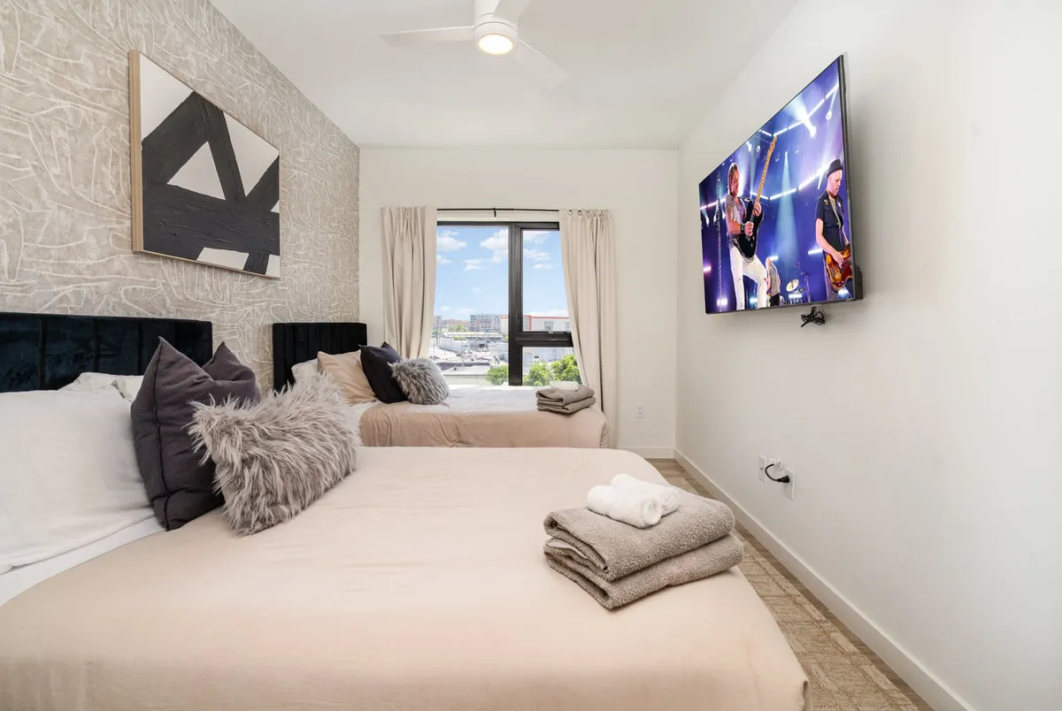 A contemporary bedroom with two beds, an oversized wall-mounted TV, abstract wall art, and a window providing natural light and a view. The room is thoughtfully arranged for comfort and flexibility, suitable for small groups or families.