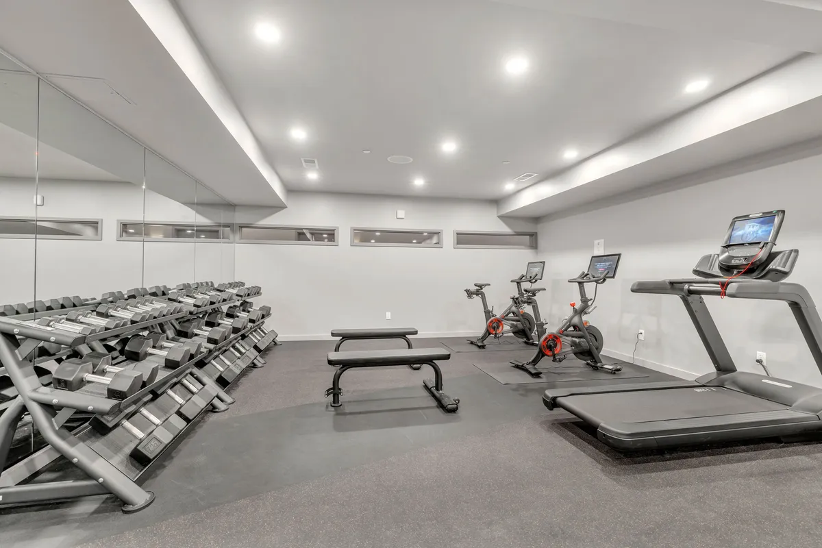 A modern, well-equipped fitness center featuring a full rack of dumbbells, treadmills, stationary bikes, and a weight bench. The space is bright, energizing, and designed for individual or group workouts.