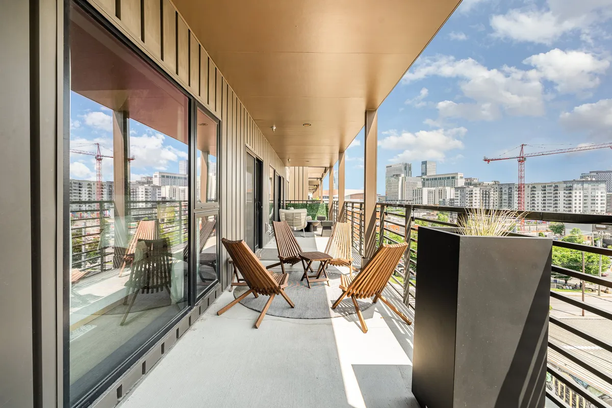 A contemporary balcony scene with wooden chairs, a planter, and a clear view of the city beyond the railing. The arrangement suggests a relaxed, social atmosphere suited for enjoying city sunsets or casual games.