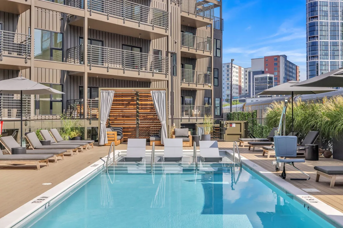 A sparkling outdoor swimming pool surrounded by lounge chairs and modern building facades. The pool area is designed for both relaxation and social gatherings, with ample space for sunbathing and enjoying the outdoors. Lush landscaping adds to the upscale atmosphere, while the building’s exterior hints at contemporary urban living.