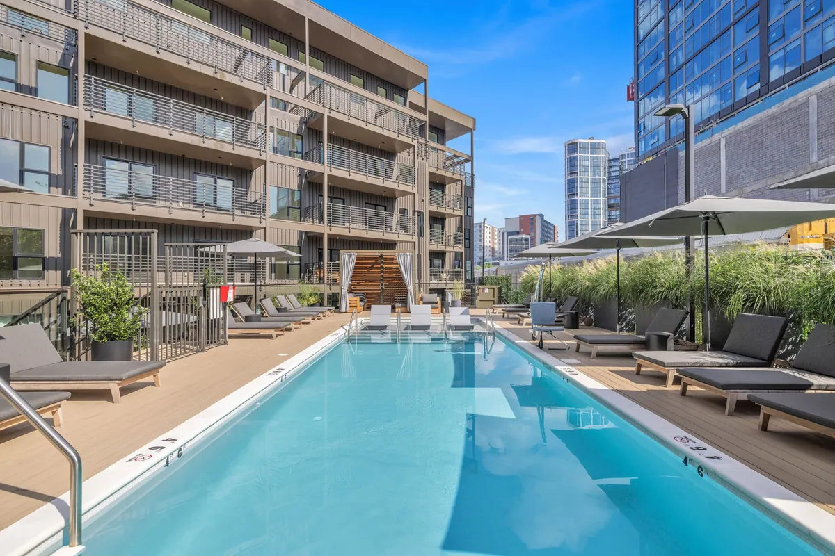 A vibrant exterior view of the resort-style swimming pool surrounded by comfortable lounge chairs and lush greenery. The pool is framed by the building facade, and the scene suggests a relaxing atmosphere perfect for sunbathing, socializing, and outdoor enjoyment.