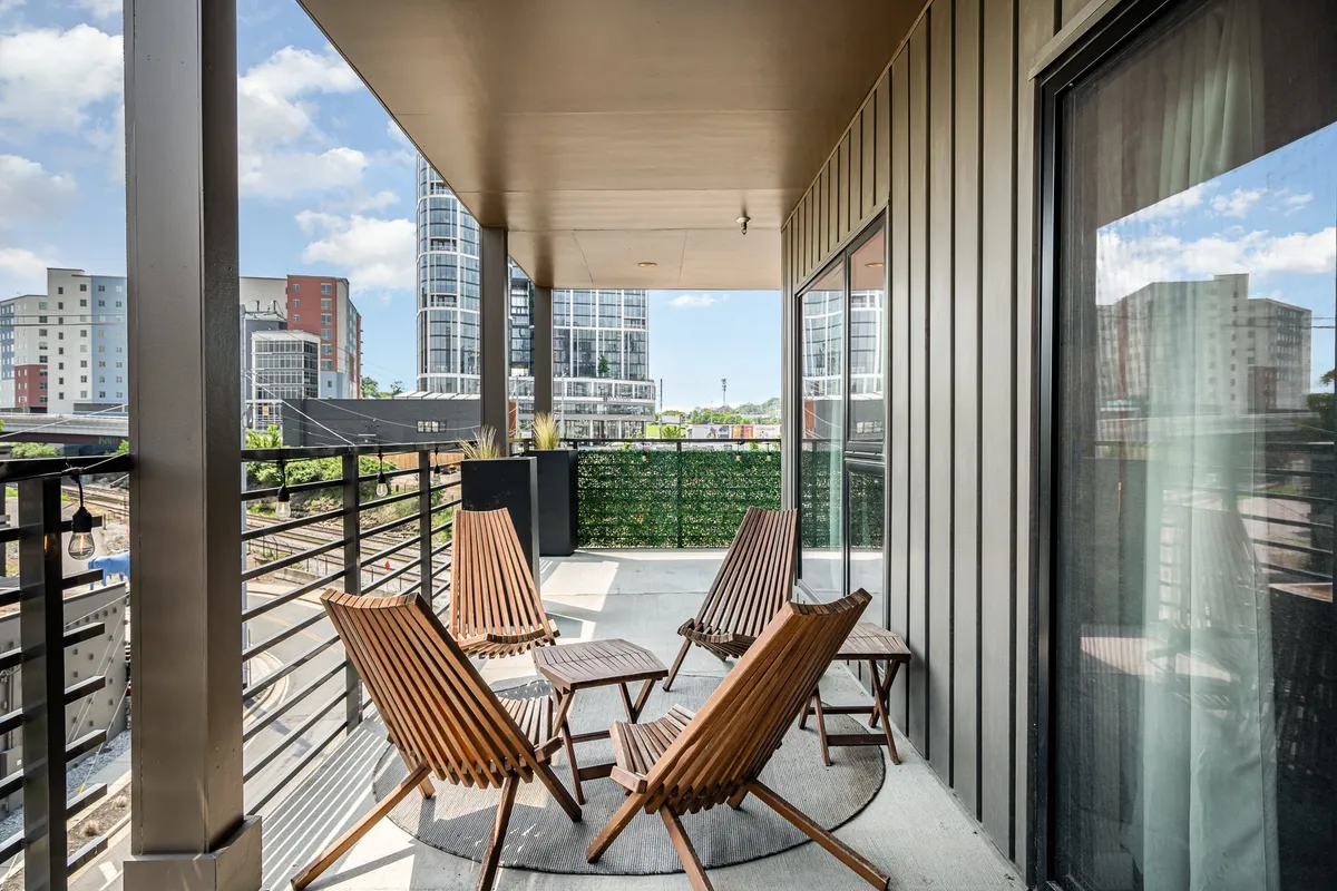 A balcony or patio area featuring wooden chairs, a planter box, and railings overlooking the cityscape. The setting invites residents or guests to relax, socialize, or enjoy quiet moments while taking in panoramic urban views—especially during sunrise or sunset.