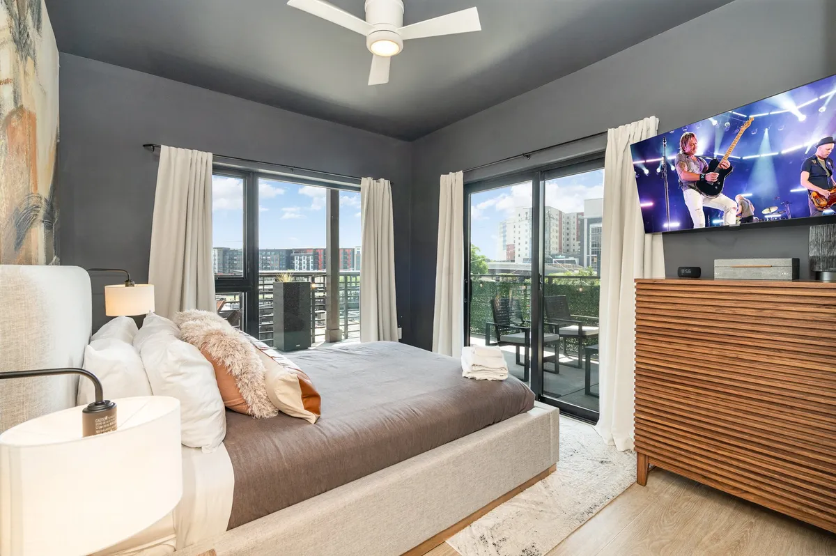 A bright, comfortable bedroom featuring a plush bed with soft gray bedding, a large window offering city views, a modern television displaying a performance, and a wooden dresser. The space feels inviting and ideal for relaxation or enjoying entertainment from bed.