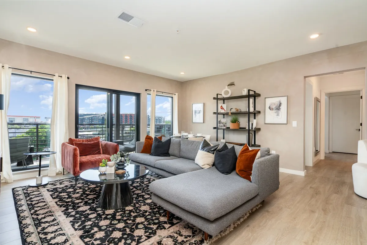 A spacious, modern living room with a plush sectional sofa, stylish coffee table, and tastefully decorated wall shelves. Sunlight streams in through large windows, offering an inviting view and creating a warm, cozy ambiance perfect for relaxation or social gatherings.