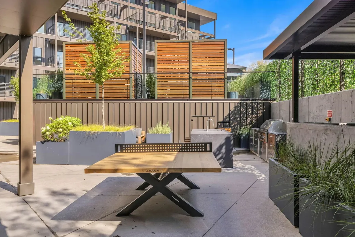 A stylish outdoor dining and grilling area with a wooden table, grill station, lush green plants, and privacy screens. The setup is ideal for barbecues, social gatherings, and relaxed community events.
